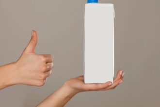 Mãos segurando uma caixa de leite longa vida com tampa azul enquanto outra mão faz sinal de positivo, em fundo neutro, representando reciclagem e reaproveitamento de embalagens.