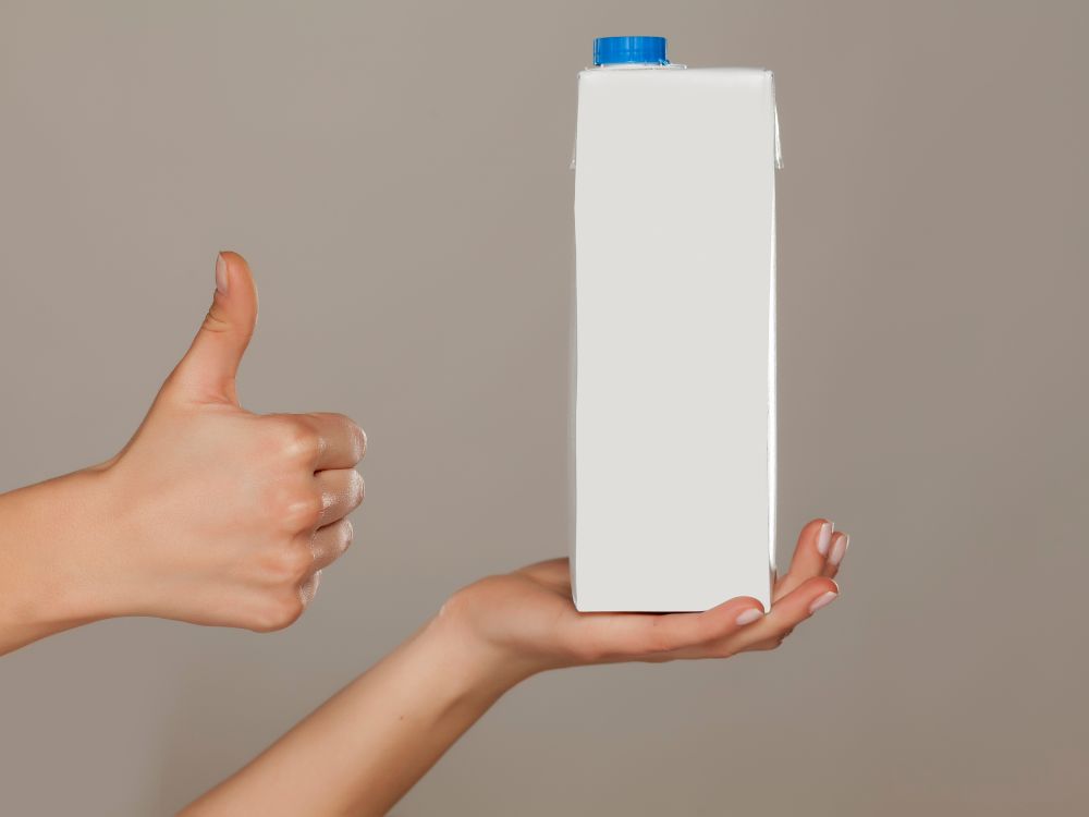 Mãos segurando uma caixa de leite longa vida com tampa azul enquanto outra mão faz sinal de positivo, em fundo neutro, representando reciclagem e reaproveitamento de embalagens.