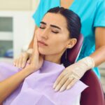 Paciente sentada em cadeira odontológica com expressão de dor, segurando a bochecha enquanto profissional prepara atendimento em consultório dentário.