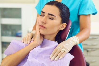Paciente sentada em cadeira odontológica com expressão de dor, segurando a bochecha enquanto profissional prepara atendimento em consultório dentário.