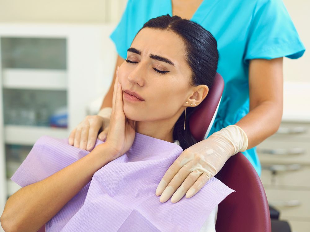 Paciente sentada em cadeira odontológica com expressão de dor, segurando a bochecha enquanto profissional prepara atendimento em consultório dentário.